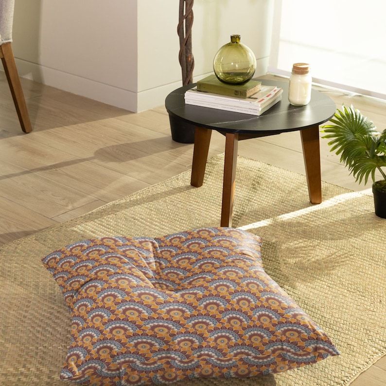 May include: A square floor cushion with a geometric pattern in shades of brown, orange, blue, and white. The cushion is sitting on a woven rug in a living room setting.