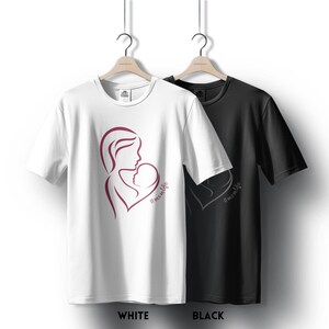 May include: Two t-shirts, one white and one black, with a design of a mother holding a baby in a heart shape. The design is in burgundy and the text below reads "#momlife".