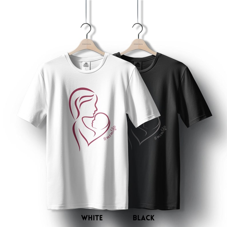 May include: Two t-shirts, one white and one black, with a design of a mother holding a baby in a heart shape. The design is in burgundy and the text below reads "#momlife".