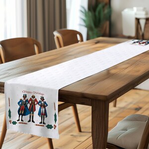 May include: White table runner with a repeating dot pattern and a festive design. The design features three toy soldier figures in blue and red coats, with the text "Happy Holidays" in red. Holly and berry accents frame the design.