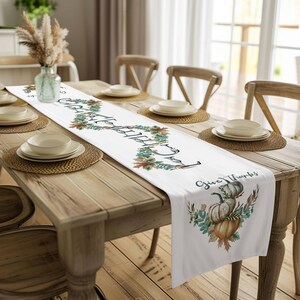 Op de afbeelding: Witte tafelloper met de woorden "Gather" en "Give Thanks" erop gedrukt. De loper heeft een bloemen- en pompoenontwerp in groen, bruin en oranje. De loper ligt op een houten tafel.