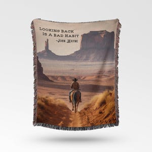 May include: A woven throw blanket featuring a quote by John Wayne: "Looking back is a bad habit." The image depicts a cowboy riding a horse through a desert landscape with a large rock formation in the background.