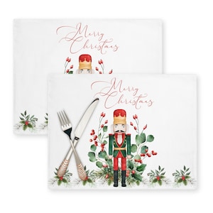 Op de afbeelding: Twee witte placemats met een aquarel-illustratie van een notenkraker, rode bessen en groen blad. De placemats hebben de tekst "Merry Christmas" in rode cursieve letters.