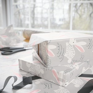 May include: Two gift boxes wrapped in gray wrapping paper with a white bunny pattern. The boxes are stacked on top of each other. The top box is partially unwrapped, revealing a white box inside. A black ribbon is on the table next to the boxes.