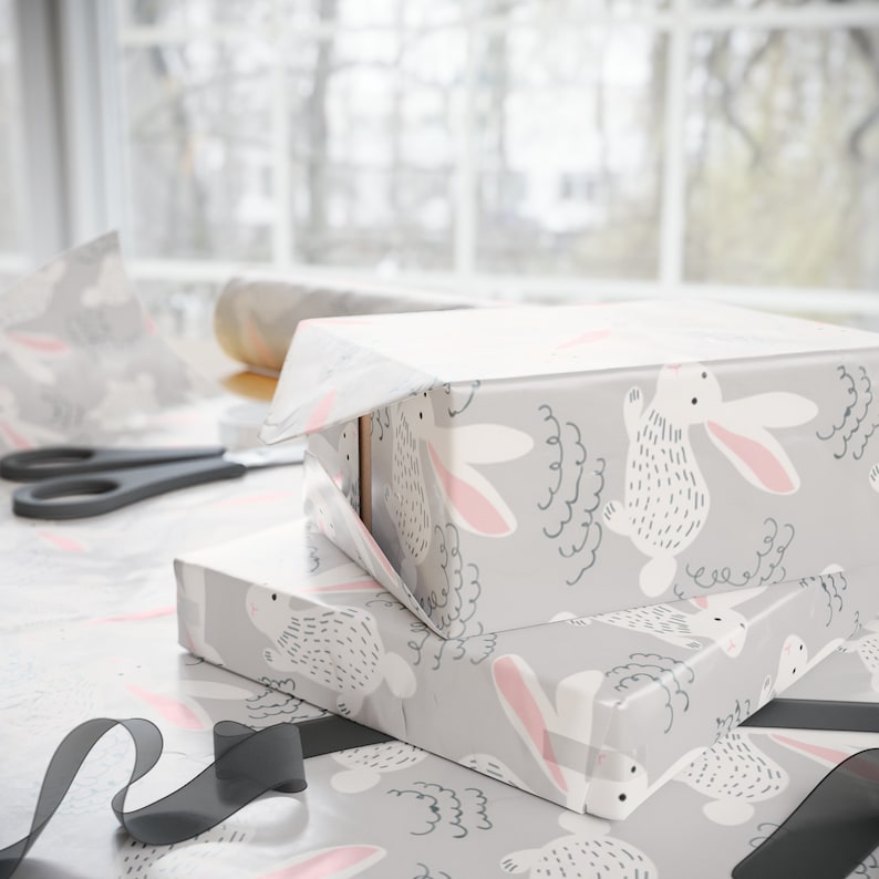 May include: Two gift boxes wrapped in gray wrapping paper with a white bunny pattern. The boxes are stacked on top of each other. The top box is partially unwrapped, revealing a white box inside. A black ribbon is on the table next to the boxes.