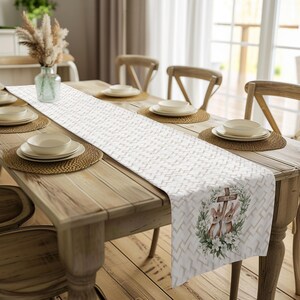 May include: A white table runner with a geometric pattern and a floral design featuring two bunnies and a cross. The runner is on a wooden table with two white plates and bowls on each side.