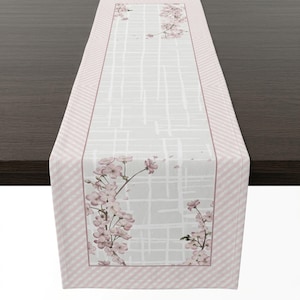 May include: A rectangular table runner with a floral design. The center features a light gray background with a white grid pattern and pink cherry blossom branches. The border has a pink and white striped pattern.