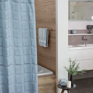 May include: A light blue shower curtain with a geometric pattern hangs in a bathroom with a white bathtub, a towel rack with a towel, and a small table with a plant and a candle.