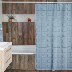 May include: A blue shower curtain with a geometric pattern of squares. The curtain is hanging in a bathroom with a white bathtub and wood-look tile.