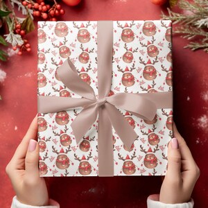 Op de afbeelding: Een ingepakt cadeau met een feestelijk rendierpatroon op wit papier. De rendieren dragen rode sjaals en mutsen. Een lichtbruin lint is om het cadeau gebonden. De achtergrond is effen rood met decoratieve elementen.