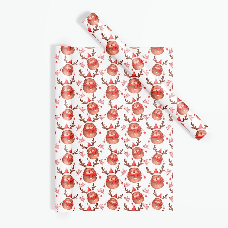 Op de afbeelding: Wit cadeaupapier met een herhalend patroon van cartoon rendieren met rode sjaals en kerstmutsen. De rendieren hebben bruine geweien en zijn omringd door rode zuurstokken. Een rol bijpassend papier is zichtbaar.