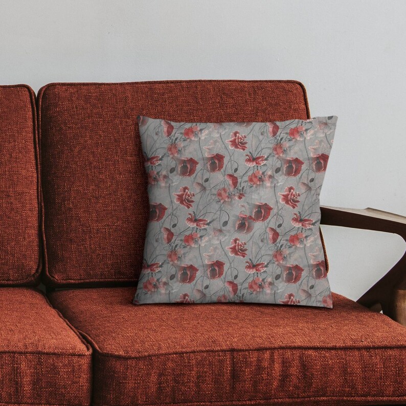 May include: Square decorative pillow with a floral pattern of red poppies on a gray background. The pillow is placed on a rust-colored sofa, creating a contrast of colors. The design features detailed botanical elements.