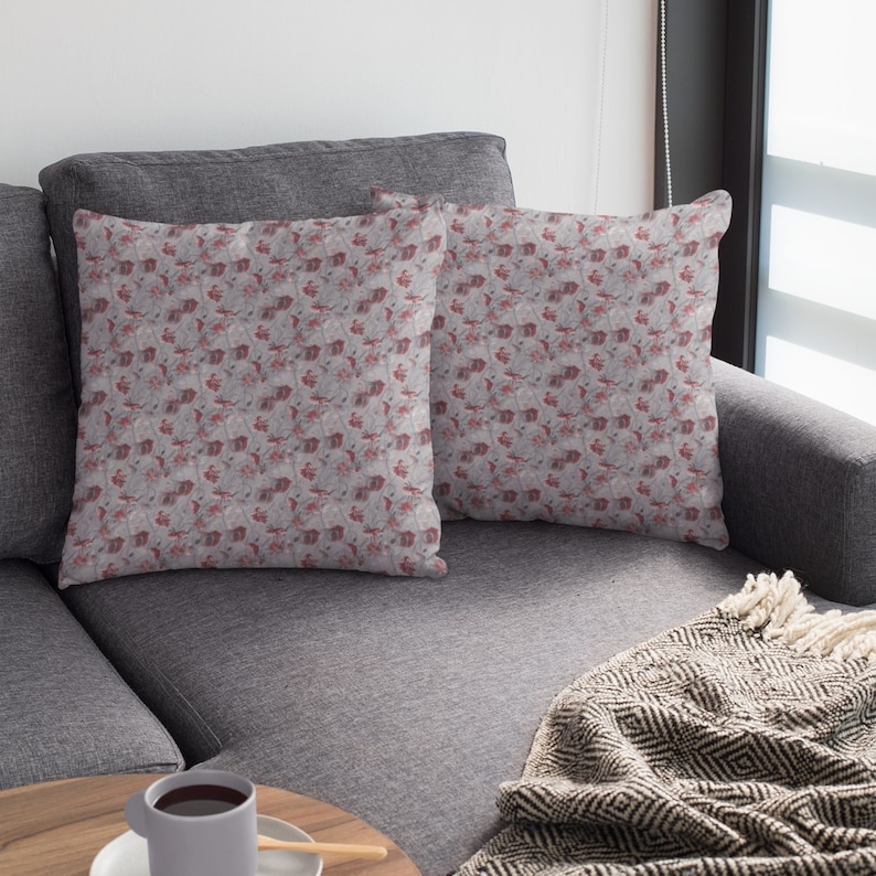 May include: Two square throw pillows with a repeating pattern of red and white floral designs on a gray background. The pillows are placed on a gray sofa, with a blanket and a cup of coffee nearby.