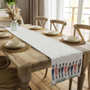 May include: White table runner with a festive design of nutcracker figurines in red, blue, and green. The runner is placed on a wooden table with place settings and a vase of dried flowers.