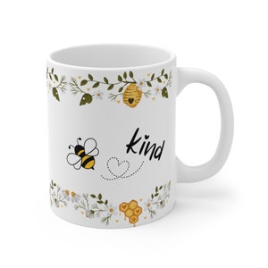 May include: White ceramic mug with a bee and honeycomb design. The mug has a black floral border and the word "kind" written in black.