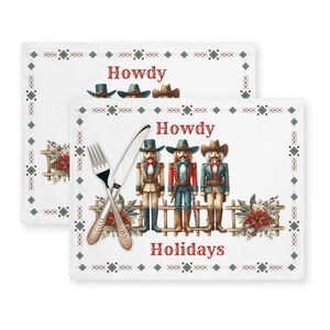 Op de afbeelding: Twee witte placemats met een groene en witte rand. De bovenste placemat heeft het woord "Howdy" in rode tekst. De onderste placemat heeft de woorden "Howdy" en "Holidays" in rode tekst. Drie notenkrakerfiguren met cowboyhoeden en geweren staan op een hek. De figuren zijn rood, blauw en groen.