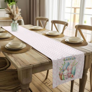May include: A pink and white chevron table runner with a bunny rabbit and Easter eggs design. The text "Happy Easter" is printed on the runner.