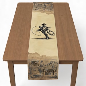 May include: A tan table runner with a western theme. The runner features a cowboy roping a steer, desert scenery, and the text "Cowboys Don't". The runner is made of a light-colored fabric and is placed on a wooden table.