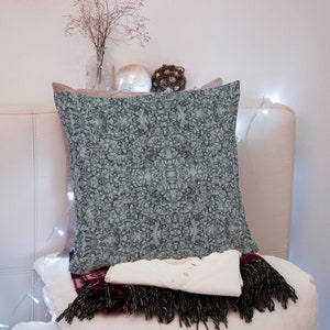 May include: A light blue throw pillow with a geometric pattern on a white couch. The pillow is sitting on top of a folded white shirt and a burgundy and black throw blanket.