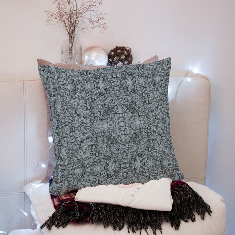 May include: A light blue throw pillow with a geometric pattern on a white couch. The pillow is sitting on top of a folded white shirt and a burgundy and black throw blanket.