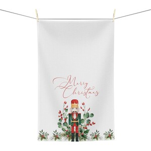 Könnte beinhalten: Weißes Küchentuch mit einer roten und grünen Aquarell-Illustration eines Nussknackers mit dem Text "Merry Christmas" in roter Kursivschrift.