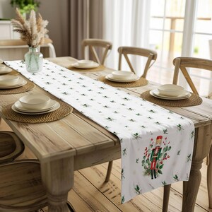 May include: White table runner with a repeating pattern of green holly leaves and red berries. A watercolor illustration of a nutcracker in a red and green uniform stands in the center of the runner.