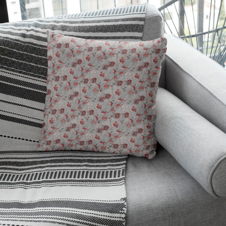 May include: A square decorative pillow with a floral pattern in shades of red and gray. The pillow is placed on a gray sofa, next to a black and white striped throw blanket. The pillow's design features a repeating pattern of stylized flowers.