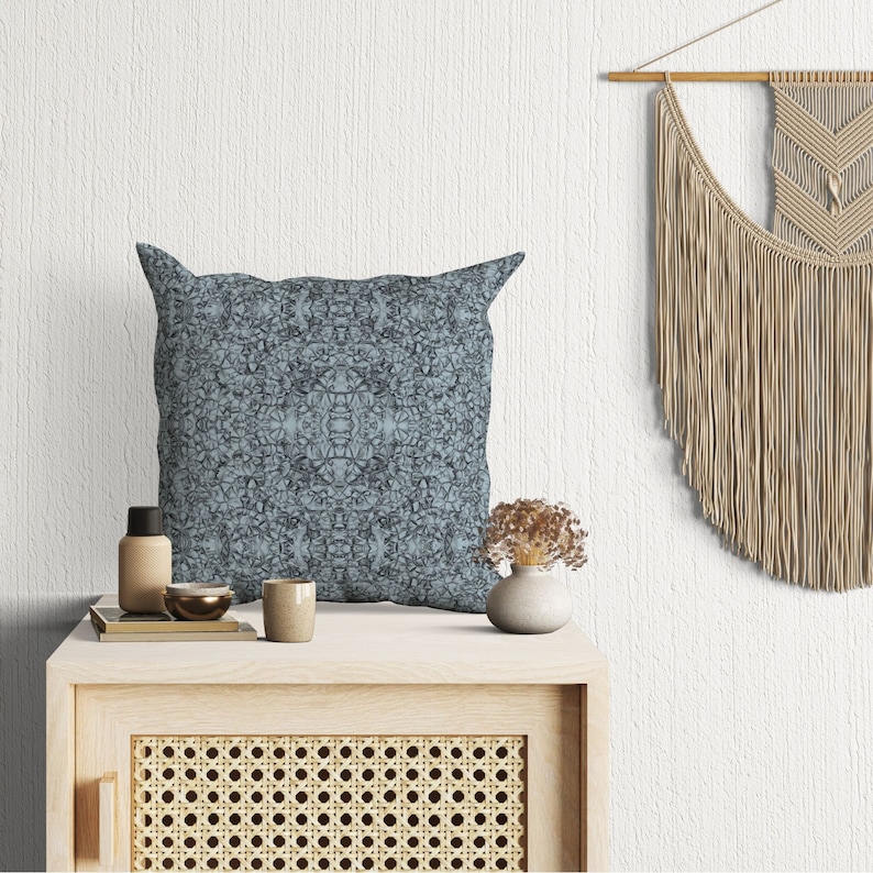 May include: A blue patterned throw pillow with a geometric design sits on a wooden table with a woven rattan front. A small vase with dried flowers and a bottle are also on the table. A macrame wall hanging is in the background.
