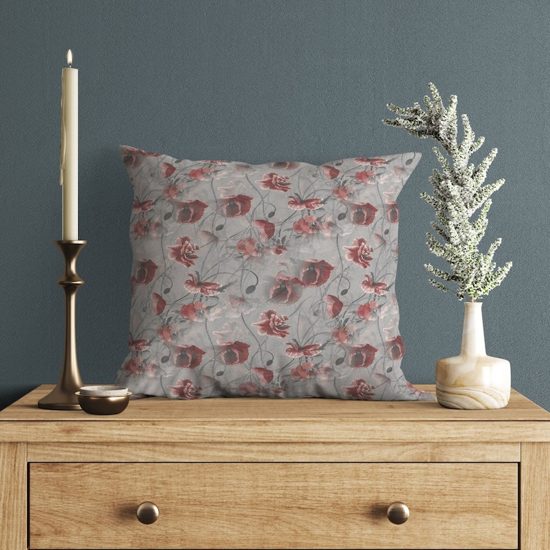 May include: A square decorative pillow with a floral pattern featuring red and pink flowers on a gray background. The pillow is placed on a wooden table, next to a lit candle in a gold candlestick and a small vase with white decorative branches.