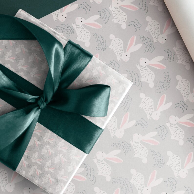 May include: A gift box wrapped in gray wrapping paper with a repeating pattern of white bunnies with pink ears. The box is tied with a dark green satin ribbon.