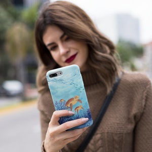 May include: A blue and white phone case with a watercolor design of sea turtles swimming in the ocean. The case features a coral reef and small fish.
