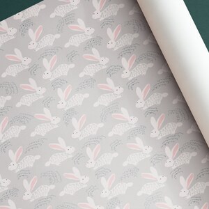 May include: A repeating pattern of white bunnies with pink ears on a light gray background. The bunnies are running and have a textured, fuzzy appearance. The background has a subtle pattern of gray lines.