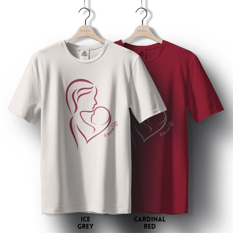 May include: Two t-shirts with a pink outline of a mother and child in a heart shape with the text #momlife. The t-shirts are on hangers. The left t-shirt is white and the right t-shirt is red.
