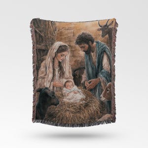 May include: A woven throw blanket featuring a detailed illustration of a nativity scene. The image depicts a person in a light-coloured head covering and a person in a blue cloak looking down at a baby lying in a bed of straw, surrounded by animals.