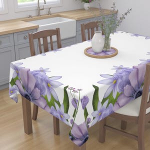 Peut inclure: Une nappe blanche avec une bordure florale violette. La nappe est sur une table en bois avec deux chaises. Il y a un vase avec des fleurs violettes et un plateau en bois sur la table.