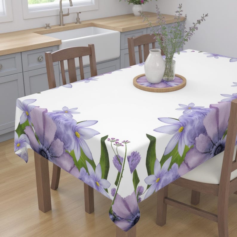 Peut inclure: Une nappe blanche avec une bordure florale violette. La nappe est sur une table en bois avec deux chaises. Il y a un vase avec des fleurs violettes et un plateau en bois sur la table.