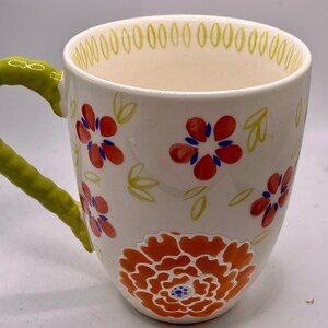 May include: White ceramic mug with a green handle and floral design. The mug features red and orange flowers with blue accents, and green leaves. The rim is decorated with small green ovals.