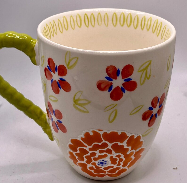 May include: White ceramic mug with a green handle and floral design. The mug features red and orange flowers with blue accents, and green leaves. The rim is decorated with small green ovals.