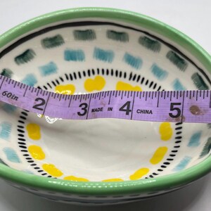 May include: A small, hand-painted ceramic bowl with a green rim and a white interior. The bowl features a decorative pattern of blue, yellow, and black accents. The bowl is approximately 6 inches in diameter.