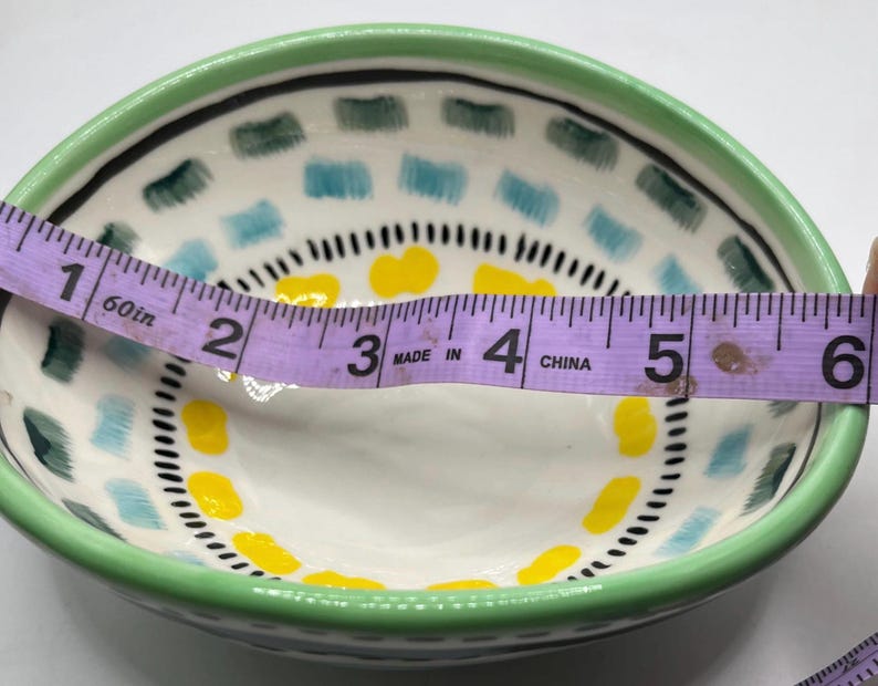 May include: A small, hand-painted ceramic bowl with a green rim and a white interior. The bowl features a decorative pattern of blue, yellow, and black accents. The bowl is approximately 6 inches in diameter.