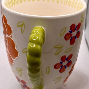 May include: White ceramic mug with a green handle and floral design. The mug features orange and red flowers with blue centers, and green leaves. The rim has a yellow dotted pattern.