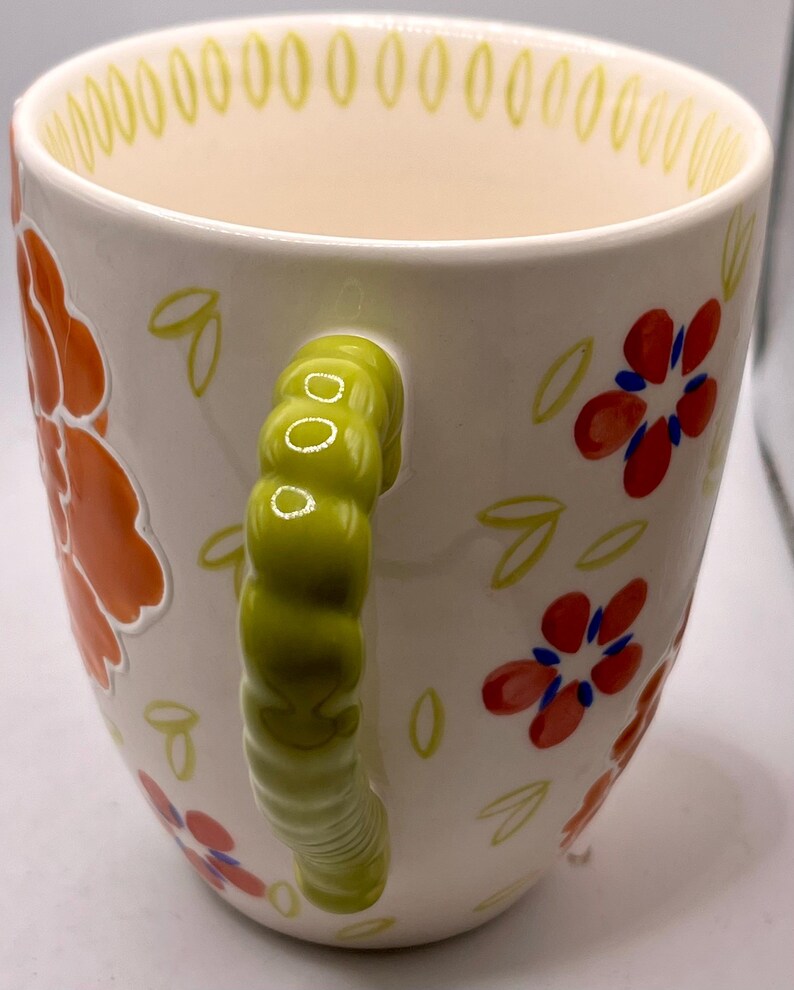 May include: White ceramic mug with a green handle and floral design. The mug features orange and red flowers with blue centers, and green leaves. The rim has a yellow dotted pattern.