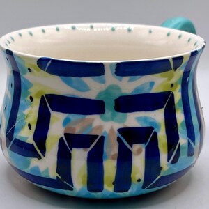 May include: A ceramic soup bowl with a turquoise handle and a white interior. The exterior features a blue, turquoise, and yellow floral design with dark blue geometric shapes. The rim has small blue dots.