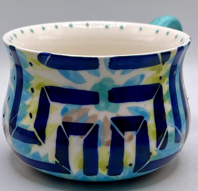May include: A ceramic soup bowl with a turquoise handle and a white interior. The exterior features a blue, turquoise, and yellow floral design with dark blue geometric shapes. The rim has small blue dots.