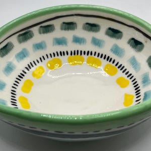 May include: A hand-painted ceramic bowl with a white interior and a green rim. The bowl is decorated with blue, green, and yellow geometric patterns. The bowl has a unique, asymmetrical shape.