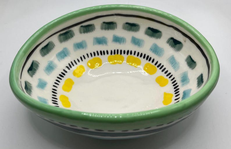 May include: A hand-painted ceramic bowl with a white interior and a green rim. The bowl is decorated with blue, green, and yellow geometric patterns. The bowl has a unique, asymmetrical shape.