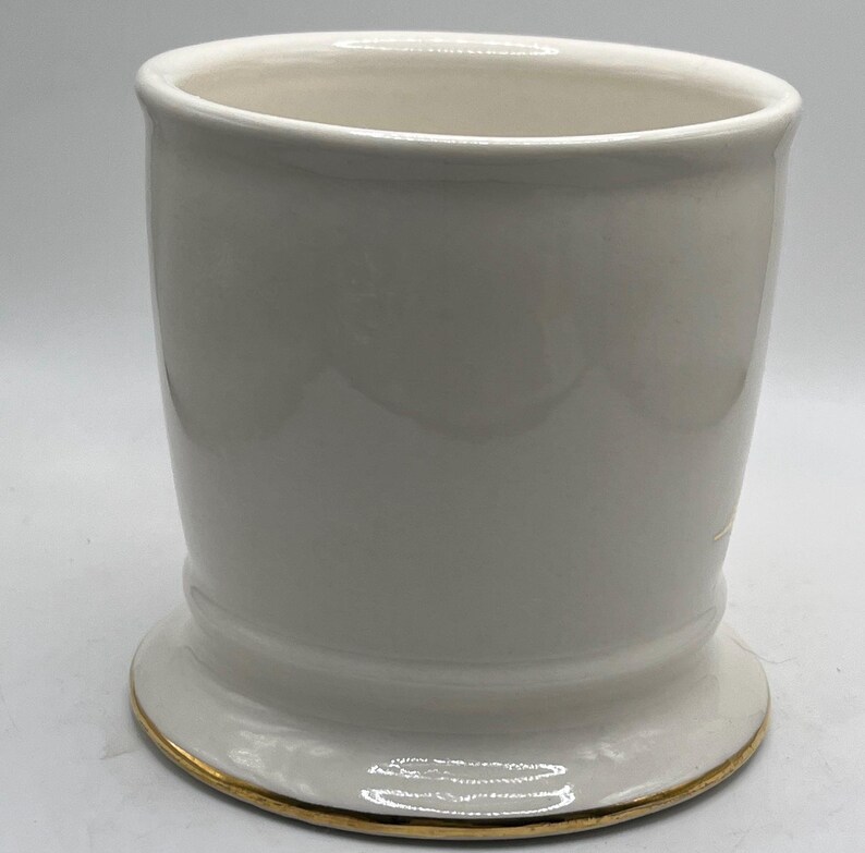 May include: White ceramic shaving mug with a gold rim. The mug has a wide base and a cylindrical shape.