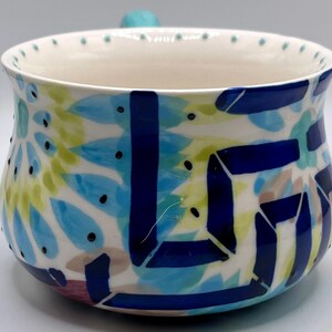 May include: A ceramic bowl with a white interior and a colorful exterior design. The exterior features blue, green, and tan abstract shapes with black dots. A small, turquoise handle is on the side.