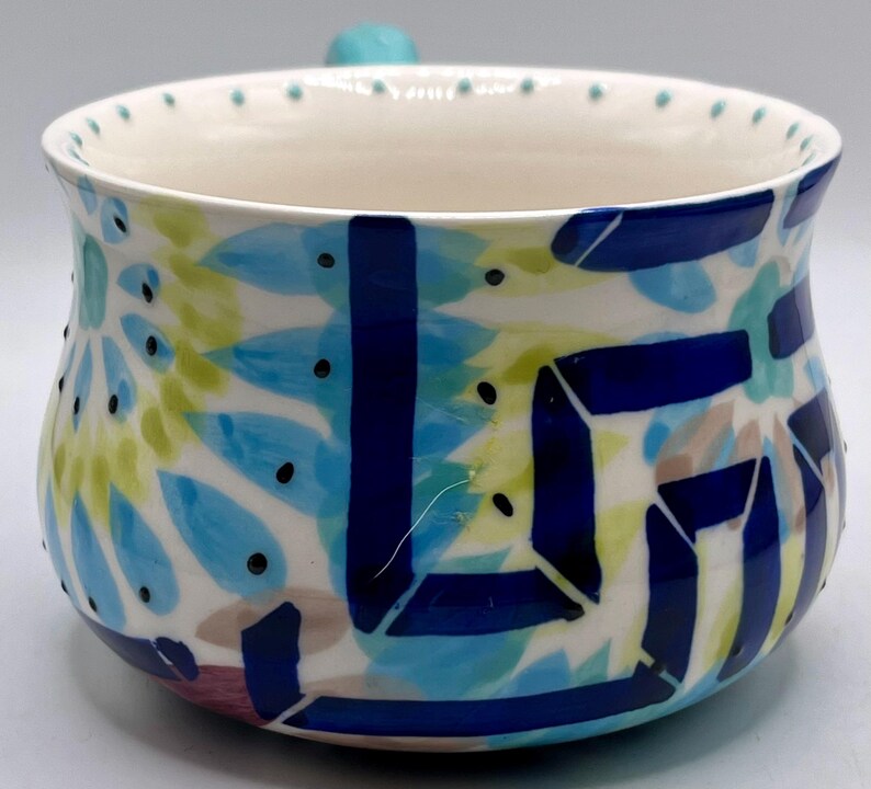 May include: A ceramic bowl with a white interior and a colorful exterior design. The exterior features blue, green, and tan abstract shapes with black dots. A small, turquoise handle is on the side.