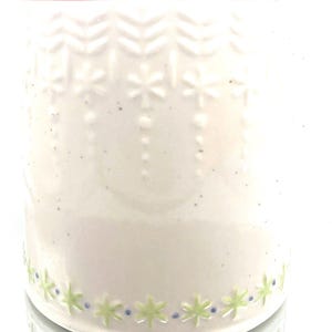 May include: A white ceramic mug with embossed snowflake and chevron patterns. The mug features a green and blue floral design around the base and a red rim. The mug is designed for beverages.